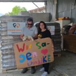 Zwei Personen halten einen Hund und ein buntes Schild mit der Aufschrift „Wir sagen DANKE“ vor gestapelten Säcken mit dem „Tierheim Tierfu“-Logo in einem Lagerraum, in dem verschiedene Vorräte herumstehen.