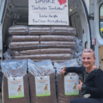 Eine lächelnde Frau zeigt vor einem mit Säcken voller Tierfutter beladenen Transporter den Daumen nach oben. Im Inneren sind Pakete mit Tierfutter gestapelt, über denen ein Dankeschön-Schild hängt, das an „Tierheim Tierfutter“ von „TierOase Aschaffenburg“ gerichtet ist.