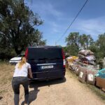 Eine Frau mit blonden Haaren steht hinter einem dunklen Transporter mit deutschem Kennzeichen, der auf einem Feldweg geparkt ist. Rechts sind Stapel aus großen Säcken und Plastikbehältern zu sehen, im Hintergrund sind unter blauem Himmel Bäume und ein weißes Auto zu erkennen.