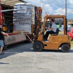 An einem sonnigen Tag bedient ein Mann einen gelben Gabelstapler und lädt eine große Palette mit verpackten Waren von einem Lkw ab. Eine weitere Person steht daneben und leitet den Vorgang.