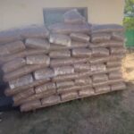 Ein großer Stapel in Plastikfolie verpackter Säcke, die möglicherweise Mulch oder Erde enthalten, ist an der Außenwand eines Gebäudes auf einer Rasenfläche aufgeschichtet.