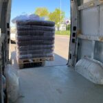 Blick aus dem Inneren eines leeren Lieferwagens auf eine große Palette, auf der Säcke mit Erde oder Mulch gestapelt sind, die in Plastikfolie eingewickelt sind und direkt hinter den offenen Türen des Lieferwagens auf der Straße stehen. Draußen sind Bäume und der blaue Himmel zu sehen.