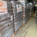 Stapel großer, in durchsichtige Plastikfolie verpackter Tierfuttersäcke stehen auf Holzpaletten in einem Lagergang. Auf den Stapeln sind Etiketten mit einem Herz- und einem Tierpfoten-Logo zu sehen.