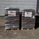 Im Freien stehen nebeneinander zwei Paletten, auf denen Säcke mit in Plastik verpacktem Tierfutter gestapelt sind. An jeder Palette ist ein Schild mit der Aufschrift „Tierheim Tierfutter“ und einem Logo mit einem Herz und einer Pfote angebracht.