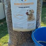 Ein großer Sack Hundefutter mit der Aufschrift „Tierheim Tierfutter“ und dem Bild eines Golden Retrievers, der draußen neben einem blauen Eimer auf grünem Gras steht.