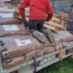 Ein älterer Mann kniet auf großen Säcken mit Tierfutter im hinteren Teil eines Anhängers und stapelt Holzpaletten. Eine weitere Person steht in der Nähe neben einem eingezäunten Bereich. Im Hintergrund sind ein grasbewachsener Hof und Holzgebäude zu sehen.