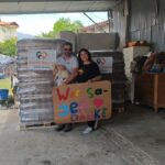 Zwei Personen stehen vor einem Stapel von Tierfuttersäcken, lächeln und halten ein buntes Schild mit der Aufschrift „Wir sagen Danke“ und Herzchen in den Händen. Eine der beiden Personen hält einen kleinen Hund im Arm. Der Schauplatz scheint ein Lagerraum zu sein.