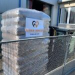 Vor einem Gebäude steht eine große, in Plastikfolie eingewickelte Palette mit Waren. Auf der Palette befindet sich ein Schild mit der Aufschrift „Tierheim Tierfutter“, einem Herz-Logo und den Worten „Spenden und Helfen“. Im Vordergrund ist ein Metallzaun zu sehen.
