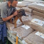 Ein Mann hält einen braunen Welpen auf Stapeln großer Hundefuttersäcke, die draußen auf einem Anhänger geladen sind; im Hintergrund sind Gras und ein Zaun zu sehen.