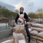 Eine Person, die eine Babytrage mit einem Kind darin trägt, steht im Freien auf gestapelten Säcken mit Pellets, im Hintergrund sind Bäume und ein bewölkter Himmel zu sehen.