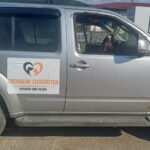 Ein silberner SUV mit einem Schild, auf dem „Tierheim Tierfutter Spenden und Helfen“ steht, sowie einem Herz mit Hunde- und Katzensilhouetten, steht an einem sonnigen Ort in der Nähe eines Zauns und einiger Gebäude.