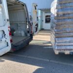 Ein offener weißer Transporter steht in der Nähe eines Gebäudeeingangs. Daneben steht eine große, in Plastikfolie eingewickelte Palette auf dem Bürgersteig, bereit zum Be- oder Entladen.