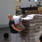 Eine Frau in Freizeitkleidung hält eine große Tüte mit Tierfutter in der Hand und steht neben einer Palette, auf der ähnliche Tüten gestapelt sind. Ein schwarzer Hund steht in der Nähe, und im Hintergrund sind Häuser und gestapeltes Brennholz zu sehen.