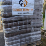 Eine große Palette, auf der Säcke mit Tierfutter gestapelt sind, die in Plastikfolie eingewickelt sind. Auf einem Schild an der Vorderseite steht „Tierheim Tierfutter Spenden und Helfen“ mit einem Herz und einem Tierlogo. Die Szene spielt im Freien.