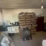 In einem schwach beleuchteten Raum stehen ein großer Stapel brauner, in Plastikfolie verpackter Pakete, eine Metallkiste, eine hängende Glühbirne, Regale mit Gläsern und verschiedene Haushaltsgegenstände.
