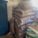In einem Raum sind mehrere große Plastiktüten, die mit etwas gefüllt sind, das wie Getreide oder ähnliches Material aussieht, hoch an einer Wand aufgestapelt. Außerdem sind ein blauer Schrank und ein Tisch mit einer gemusterten Tischdecke zu sehen.