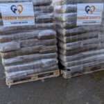 Zwei Paletten, auf denen Säcke mit Tierfutter gestapelt sind, die in Plastikfolie verpackt sind. Auf jeder Palette befindet sich ein Schild mit der Aufschrift „Tierheim Tierfutter, Spenden und Helfen“. Die Paletten stehen draußen auf einer gepflasterten Fläche, im Hintergrund sind Bäume zu sehen.