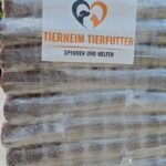 Eine Palette mit in Plastikfolie verpackten Säcken mit Tierfutter, mit einem Schild mit der Aufschrift „Tierheim Tierfutter Spenden und Helfen“ und einem Logo, das einen Hund und eine Katze in Herzform zeigt.