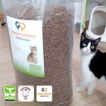 Eine schwarz-weiße Katze steht neben einem großen Sack mit Trockenfutter. Auf dem Sack ist ein Etikett mit dem Bild eines Kätzchens zu sehen. In der unteren linken Ecke sind drei Logos von Organisationen zu erkennen. Die Szene spielt in einem Innenraum.