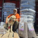 Ein Mann mit einer Mütze kniet neben einem hellfarbigen Hund, dessen Bein bandagiert ist. Sie befinden sich vor großen Stapeln von Tierfuttersäcken und Paletten, vermutlich im Inneren eines Lastwagens.