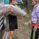 Ein Junge in schwarzen Shorts trägt in der Nähe eines Lastwagens einen großen Sack mit Holzspänen auf der Schulter, während eine Frau in einem lila Kapuzenpulli und gestreiften Hosen im Freien ihm gegenüber steht.