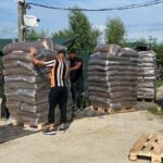 Zwei Personen stehen im Freien neben großen Stapeln von in Säcke verpackten Waren auf Holzpaletten. Eine Person richtet einen Stapel aus, während die andere neben den Paletten steht. Der Himmel ist klar, und im Hintergrund sind Bäume zu sehen.