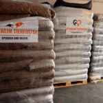 Große Stapel von in Plastikfolie verpackten Tierfutterbeuteln auf Holzpaletten, beschriftet mit „Tierheim Tierfutter“ und mit Spendenaufrufen in deutscher Sprache versehen, gelagert in einem Lagerhaus.