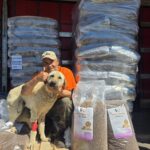 Ein Mann in einem orangefarbenen Hemd und einer orangefarbenen Mütze sitzt mit einem großen Hund vor einem Lkw, in dem Säcke mit Hundefutter gestapelt sind, und streckt den Daumen nach oben. Neben ihnen liegen zwei geöffnete Säcke mit Hundefutter auf dem Boden.