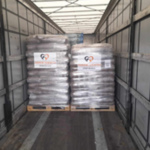 Zwei Paletten mit verpackten Säcken werden in einen leeren LKW-Anhänger geladen. Jede Palette ist mit einem gut sichtbaren Etikett versehen, auf dem ein Logo und ein Text zu sehen sind. Die Seitenwände und das Dach des Anhängers sind zu sehen.