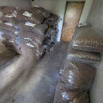 Zu sehen ist ein kleiner Raum mit Stapeln großer, durchsichtiger Plastiktüten, die mit braunen Kügelchen gefüllt sind; einige sind an den Wänden aufgestapelt, andere auf dem Boden aufgereiht, und am Ende befindet sich eine geschlossene Holztür.
