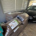 Große Säcke mit Trockenfutter für Haustiere sind neben einem schwarzen Auto in einem überdachten Bereich mit Steinboden und weißen Jalousien an den Fenstern aufgestapelt.