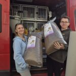 Zwei Personen stehen mit großen Säcken Hundefutter neben einem roten Transporter. Hinter ihnen stehen die Türen des Transporters offen und geben den Blick auf Tierkäfige und Zubehör im Inneren frei. Beide lächeln und sind leger gekleidet.