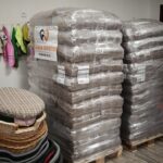 In einem Raum stehen zwei große Stapel mit in Plastikfolie verpackten Tierfutterbeuteln auf Paletten. Daneben stehen Weidenkörbe mit karierten Kissen, und an der Wand hängen Tierzubehör wie Leinen und Jacken.