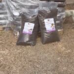 Zwei große Säcke mit Tierfutter stehen im Freien aufrecht vor einem Stapel verpackter Paletten. Ein weißer Hund läuft in der Nähe der Säcke umher, und der Boden ist mit Erde und Stroh bedeckt.