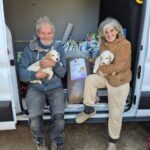 Zwei lächelnde Erwachsene sitzen auf der Rückbank eines Kleinbusses und halten jeweils einen Welpen im Arm. Zwischen ihnen steht eine große Tüte mit Hundefutter und Zubehör. Die Tür des Kleinbusses ist offen, und die Szene scheint im Freien zu spielen.