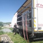 Ein Mann steht neben einem offenen Lastwagen, der mit in Plastikfolie verpackten, gestapelten Waren beladen ist. Die Seitenplane des Lastwagens ist zurückgezogen, und auf dem Rasen daneben liegen unter einem strahlend blauen Himmel Holzpaletten.