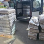 Mehrere Stapel von in Plastikfolie verpackten Pelletsäcken liegen auf Paletten und auf dem Boden neben einem offenen Transporter, wobei im Inneren des Transporters weitere Säcke zu sehen sind. Die Szene scheint in einem Lagerhaus oder einem Verladebereich zu spielen.