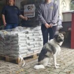 Vor einem Gebäude stehen zwei Frauen neben auf Paletten gestapelten Säcken mit Hundefutter. Eine von ihnen zeigt auf ein Schild mit einem Hundelogo. Vor ihnen sitzt ein großer Hund auf einem gepflasterten Untergrund.