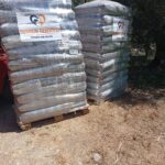 Zwei Paletten, auf denen Säcke mit in Plastikfolie verpacktem Tierfutter gestapelt sind, die mit der Aufschrift „Tierheim Tierfutter Spenden und Helfen“ versehen sind, stehen im Freien auf trockenem Gras in der Nähe einiger Bäume.
