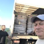Zwei Männer stehen neben einem offenen Lastwagen, der mit gestapelten Säcken oder Paketen beladen ist. Ein Mann macht im Vordergrund ein Selfie, während der andere neben der Laderampe des Lastwagens steht. Es ist hellichter Tag, und der Himmel ist klar.