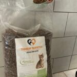 Eine Hand schöpft Trockenfutter für Katzen aus einem großen durchsichtigen Beutel mit der Aufschrift „Tierheim Tierfutter“ und dem Bild einer Katze vor einem gefliesten Hintergrund.