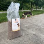 Auf einem Gehweg steht ein großer Sack Hundefutter, während ein schwarz-weißer Hund in der Nähe einer Rasenfläche mit Bäumen im Hintergrund Trockenfutter vom Boden frisst.