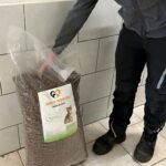 Eine Person in dunkler Kleidung steht in einem Raum und hält neben einer gefliesten Wand einen großen Sack Katzenfutter mit der Aufschrift „Tierheim Tierfutter“ und einem Katzenbild in der Hand.