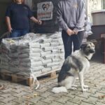 Zwei lächelnde Frauen stehen draußen neben gestapelten Säcken mit Tierfutter auf Holzpaletten. Vor ihnen sitzt ein großer Hund auf einem Kopfsteinpflasterweg. Die Kulisse scheint ein Tierheim oder eine Tierschutzorganisation zu sein.