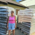 Eine Frau in einem rosa Tanktop und blauen Shorts steht zwischen zwei großen Paletten mit in Schrumpffolie verpackten Säcken.