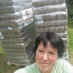 Eine Frau macht draußen ein Selfie vor zwei großen Paletten, auf denen verpackte Säcke gestapelt sind, die mit einem Herz-Logo und einem Schriftzug versehen sind. Im Hintergrund sind Gras und Grünpflanzen zu sehen.