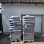 Zwei Paletten mit in Schrumpffolie verpackten Säcken voller Holzpellets vor einem Gebäude mit einem weißen Garagentor.