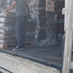 Ein Mann in einem gestreiften Hemd und mit einer Mütze steht in einem Lastwagen, in dem auf Paletten gestapelte, in Plastikfolie eingewickelte Säcke liegen. Die Seite des Lastwagens ist offen, und auf einem der Säcke ist ein Logo mit einem Hund zu sehen. Darunter ist der Kiesboden zu sehen.