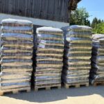 Vier Paletten mit in Schrumpffolie verpackten Säcken voller Erde, die auf einem gepflasterten Untergrund an einer Holzscheune gestapelt sind.