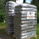 Auf dem Rasen stehen zwei hohe Stapel aus in Plastikfolie verpackten Säcken auf Holzpaletten. An jedem Stapel befindet sich ein Schild mit einem Herz-Logo und der Aufschrift Tierheim Tierfutter auf der Vorderseite. Im Hintergrund sind Bäume und Grünflächen zu sehen.